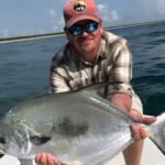 A photo of a man in a pair of sunglasses and a cap holds up a saltwater fish.