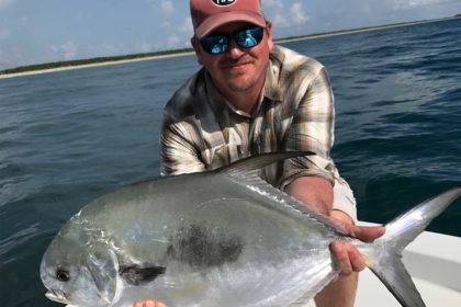 A photo of a man in a pair of sunglasses and a cap holds up a saltwater fish.