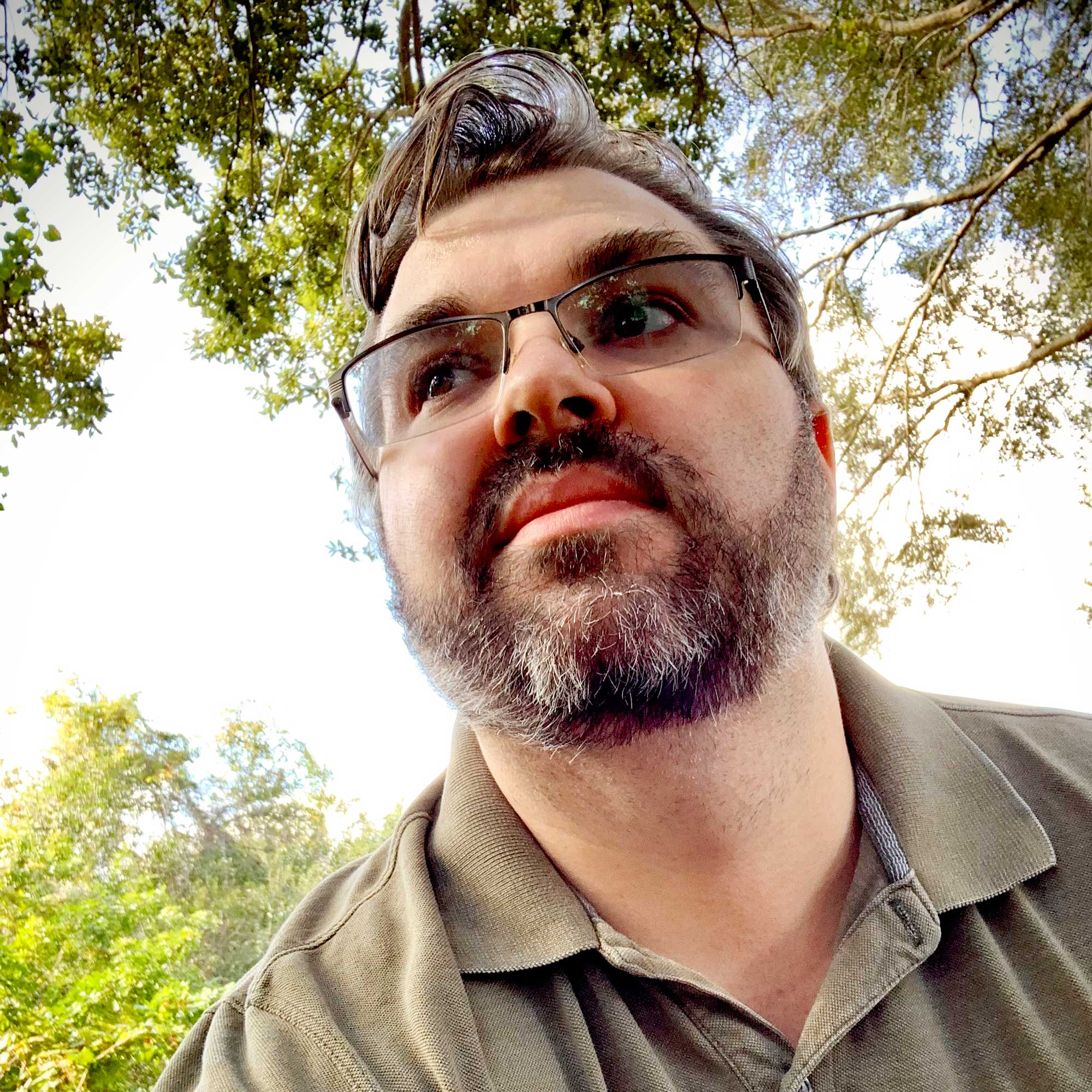 Photo of a bearded man wearing glasses in front of a clear blue sky and tree foliage.